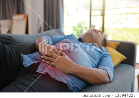 Overweight man lying on sofa holding stomach with red inflamed spot from abdominal pain. Concept of indigestion, gastritis, food poisoning, stomachache, and unhealthy lifestyle in living room. Overweight man lying on sofa holding stomach with red inflamed spot from abdominal pain. Concept of indigestion, gastritis, food poisoning, stomachache, and unhealthy lifestyle in living room. 135944223