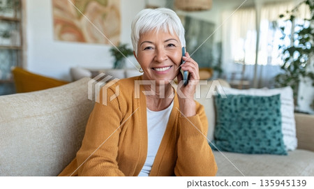 Smiling senior woman with short silver hair enjoying a phone call on a smartphone while sitting comfortably in a bright living room with cozy decor 135945139