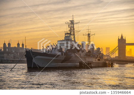 HMS Belfast sits on the River Thames in London during the morning. The iconic Tower Bridge stands in the background as the sun rises, illuminating the city. 135946664