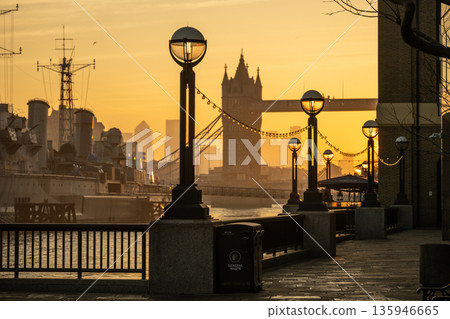The sun rises over the Thames River in London. Buildings stand in the distance while street lamps line the walkway. Boats quietly float on the water as a new day begins. 135946665