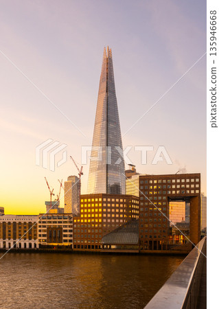 Sunlight shines on The Shard as it rises above the buildings of London. The city prepares for a new day with construction activity visible in the scene. 135946668