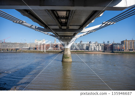 The underside of the Millennium Bridge in London. The bridge spans the River Thames. Buildings and structures line the riverbank on both sides. The underside of the Millennium Bridge in London. The bridge spans the River Thames. Buildings and structures line the riverbank on both sides. 135946687