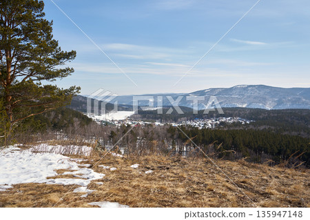 Hilltop view with dry grass and partial snow with a winter village, evergreen forest, and mountains under a blue winter sky for nature travel. Hilltop view with dry grass and partial snow with a winter village, evergreen forest, and mountains under a blue winter sky for nature travel. 135947148