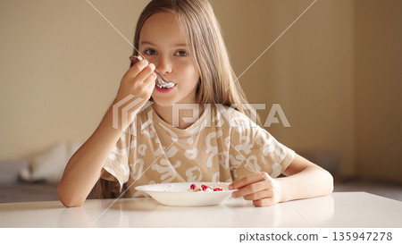 Happy cute girl eating cottage cheese with fresh berries at a table, enjoying a healthy snack indoors with natural light and a calm atmosphere, looking at camera 135947278
