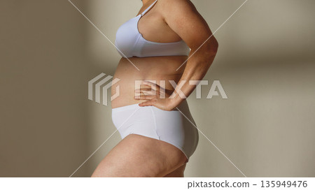 Older woman wearing white lingerie standing on studio background. Older woman wearing white lingerie standing on studio background. 135949476