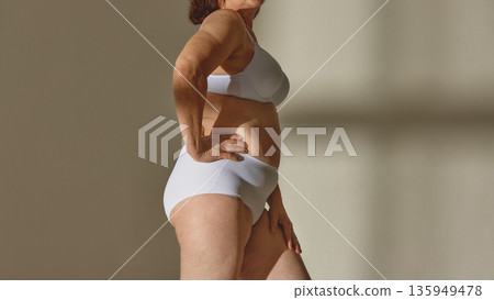 Confident mature woman in white lingerie standing in studio. Confident mature woman in white lingerie standing in studio. 135949478