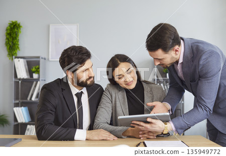 Office people, couple, colleagues gathered around desk looking attentively at tablet computer Office people, couple, colleagues gathered around desk looking attentively at tablet computer 135949772
