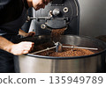 Young male employee controlling the process of pouring freshly roasted beans into a drum.  Man working at an industrial roasting machine. 135949789