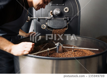 Young male employee controlling the process of pouring freshly roasted beans into a drum. Man working at an industrial roasting machine. Young male employee controlling the process of pouring freshly roasted beans into a drum. Man working at an industrial roasting machine. 135949789