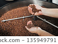 Close-up of male hands holding freshly roasted coffee beans while checking roasting quality. Drum of a professional coffee roasting machine filled with beans in the background 135949791