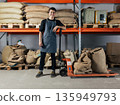 Smiling warehouse worker leaning on a manual forklift trolley in a storage facility. Young cheerful man wearing an apron relaxes during a warehouse shift and looks at the camera. 135949793