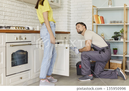 Man plumber fixing broken pipe in modern kitchen and talking with female client at home. 135950996