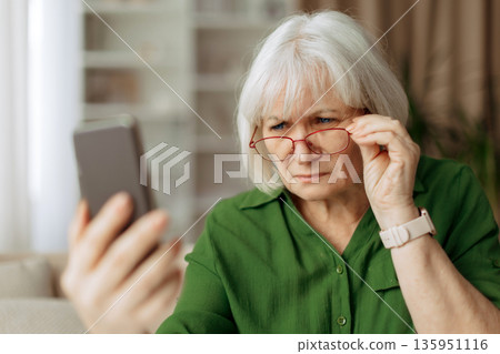 Senior woman squinting eyes and adjusting red glasses while looking at smartphone Senior woman squinting eyes and adjusting red glasses while looking at smartphone 135951116