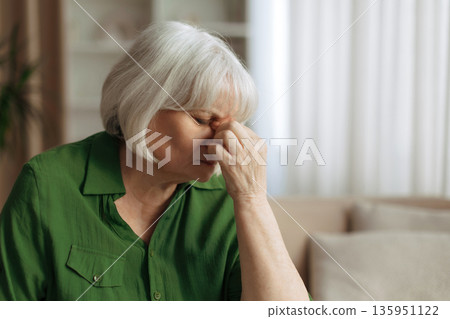 Elderly woman rubbing nose and closing eyes at home, suffering from headache Elderly woman rubbing nose and closing eyes at home, suffering from headache 135951122