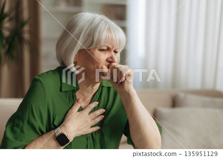 Elderly caucasian woman coughing while holding chest at home, experiencing respiratory illness Elderly caucasian woman coughing while holding chest at home, experiencing respiratory illness 135951129
