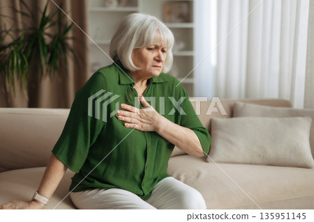 Senior caucasian woman leaning forward and touching chest at home Senior caucasian woman leaning forward and touching chest at home 135951145
