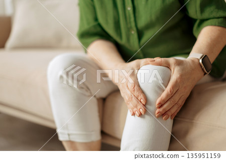 Elderly caucasian woman holding knee while sitting on sofa at home Elderly caucasian woman holding knee while sitting on sofa at home 135951159