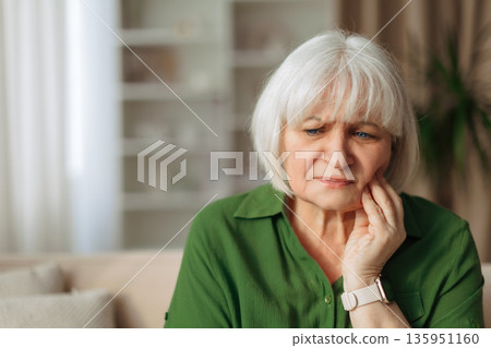 Elderly woman touching cheek with worried expression at home, feeling tooth pain Elderly woman touching cheek with worried expression at home, feeling tooth pain 135951160