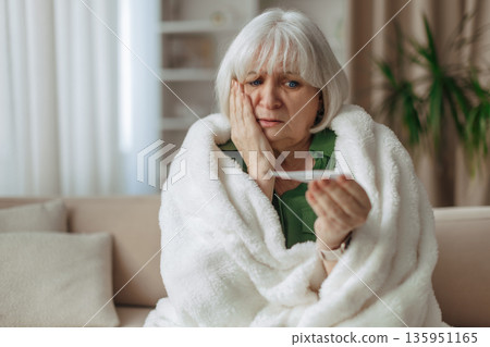 Elderly caucasian woman looking worried while holding thermometer at home Elderly caucasian woman looking worried while holding thermometer at home 135951165