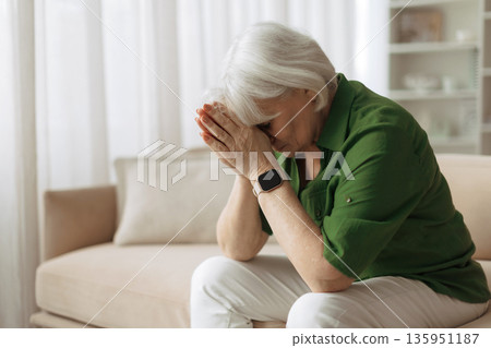 Elderly woman sitting with hands covering face at home, experiencing sadness Elderly woman sitting with hands covering face at home, experiencing sadness 135951187