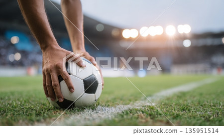 Striker prepares for soccer penalty kick, hands gripping ball on grass field, low-angle view highlighting stadium lights and atmosphere 135951514