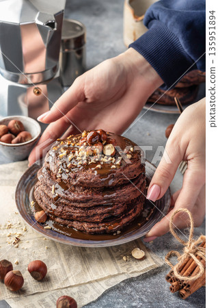 Womans hands put a plate of yummy chocolate pancakes topped with nuts and drizzled with chocolate syrup on a table. Cozy breakfast and coffee on gray background Womans hands put a plate of yummy chocolate pancakes topped with nuts and drizzled with chocolate syrup on a table. Cozy breakfast and coffee on gray background 135951894