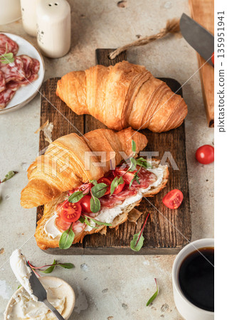 Croissant sandwich with salami, sausage, cherry tomatoes, soft cheese and chard micro greens on wooden board kitchen table background for savory healthy breakfast Croissant sandwich with salami, sausage, cherry tomatoes, soft cheese and chard micro greens on wooden board kitchen table background for savory healthy breakfast 135951941