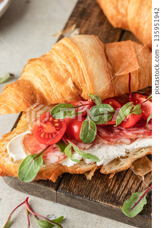 Croissant sandwich with salami, sausage, cherry tomatoes, soft cheese and chard micro greens on wooden board close up for savory healthy breakfast Croissant sandwich with salami, sausage, cherry tomatoes, soft cheese and chard micro greens on wooden board close up for savory healthy breakfast 135951942