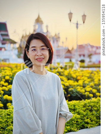 Healthy middle-aged woman stands in front of a sunlit garden of yellow flowers with ornate temple architecture softly blurred in the background at golden hour. Healthy middle-aged woman stands in front of a sunlit garden of yellow flowers with ornate temple architecture softly blurred in the background at golden hour. 135954118