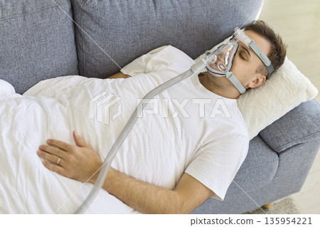 Man resting in bed with oxygen mask, receiving breathing support therapy at home Man resting in bed with oxygen mask, receiving breathing support therapy at home 135954221