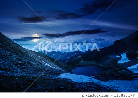 mountain lake in alpine summer landscape at night. beautiful view surrounded with clouds on a dark sky in full moon light. fresh water, snow and rocky slopes. mysterious remote fantasy highlands mountain lake in alpine summer landscape at night. beautiful view surrounded with clouds on a dark sky in full moon light. fresh water, snow and rocky slopes. mysterious remote fantasy highlands 135955177