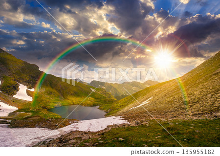 mountain lake in alpine summer landscape at sunset. beautiful view surrounded with clouds on a blue sky in evening light. fresh water, snow and rocky slopes. remote highlands adventures under rainbow 135955182
