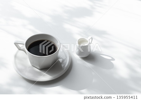 白色背景上的黑色咖啡和牛奶壺。陽光透過樹木和空白處投下斑駁的光影。 白色背景上的黑色咖啡和牛奶壺。陽光透過樹木和空白處投下斑駁的光影。 135955411