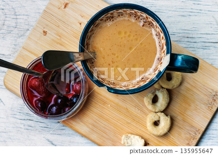 Top view of a coffee cup on a wooden board with cookies, butter, and an open jar of wildberry jam with a spoon, on a light tabletop with copy space. Top view of a coffee cup on a wooden board with cookies, butter, and an open jar of wildberry jam with a spoon, on a light tabletop with copy space. 135955647
