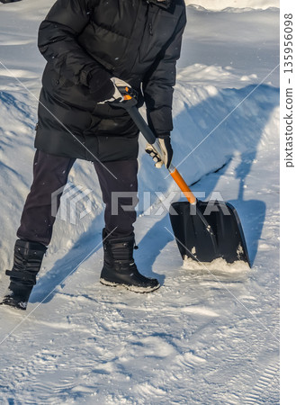 A person in a black jacket shovels snow on a sunny day after a blizzard. A person in a black jacket shovels snow on a sunny day after a blizzard. 135956098