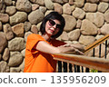 Elegant woman in orange dress standing against rustic stone wall, wearing sunglasses outdoors 135956209