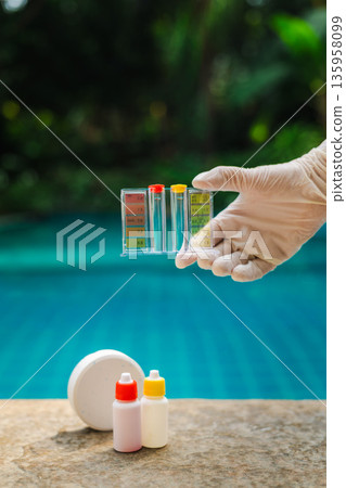 Gloved hand holding pool water test kit over swimming pool with chlorine tablet and reagent bottles on stone edge, water quality control and sanitation concept 135958099