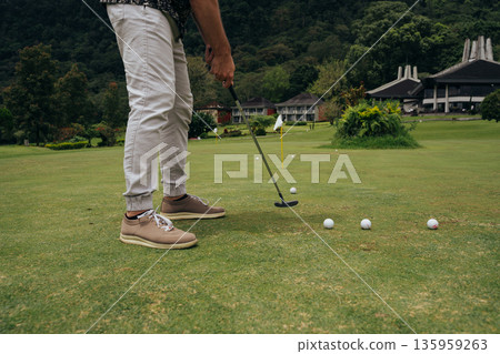 A golfer perfecting their putt on a lush green course surrounded by scenic mountains in the afternoon light 135959263