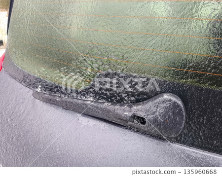 Close-up of car rear window wiper frozen in place by thick ice and frost, demonstrating harsh winter conditions affecting vehicle functionality. Close-up of car rear window wiper frozen in place by thick ice and frost, demonstrating harsh winter conditions affecting vehicle functionality. 135960668