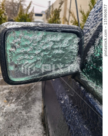 Close-up of icy green car side mirror encrusted in winter frost showing textured surface, frozen droplets and reduced clarity through glazed frozen glass. 135960677