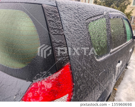 Rear section of dark vehicle with icy coating on windows and taillight, showing heavy frost accumulation and limited visibility during freezing winter weather. Rear section of dark vehicle with icy coating on windows and taillight, showing heavy frost accumulation and limited visibility during freezing winter weather. 135960698