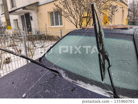 Black car windshield with raised wiper blade against frozen glass showing ice layer beneath winter frost and textured haze on vehicle surface. Black car windshield with raised wiper blade against frozen glass showing ice layer beneath winter frost and textured haze on vehicle surface. 135960701