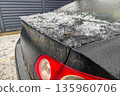 Partial view of black car rear with snowy roof and icy buildup beside red taillight, capturing winter street parked vehicle conditions with frost layers. 135960706