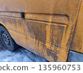 Dirty yellow van with road grime streaks parked on snowy surface, showing contrast between muddy residue and bright paint during winter weather conditions. 135960753