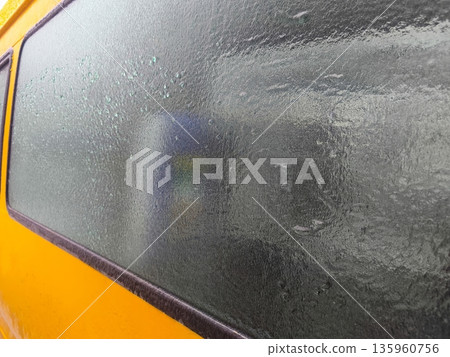 Frozen condensation completely covering yellow van window, creating textured icy surface and blurred view of interior objects through thick winter frost. 135960756