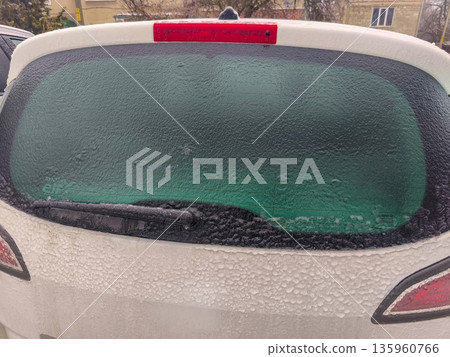 Frozen rear windshield of white vehicle with greenish tint and complete ice coverage, creating opaque appearance due to heavy winter frost layer. Frozen rear windshield of white vehicle with greenish tint and complete ice coverage, creating opaque appearance due to heavy winter frost layer. 135960766