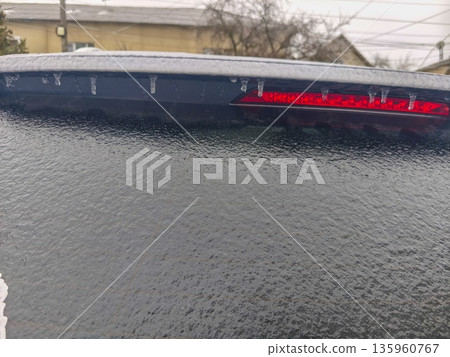 Frozen rear windshield with thin ice layer and red brake light barely visible beneath frost, showing winter driving conditions and reduced visibility. Frozen rear windshield with thin ice layer and red brake light barely visible beneath frost, showing winter driving conditions and reduced visibility. 135960767