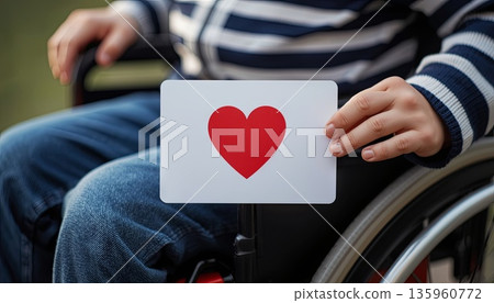 A disabled boy sits in a wheelchair and holds a Valentine's Day card. The concept is about Valentine's Day, inclusivity, love for others, and caring. A disabled boy sits in a wheelchair and holds a Valentine's Day card. The concept is about Valentine's Day, inclusivity, love for others, and caring. 135960772