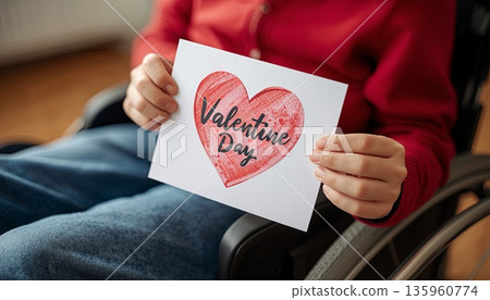A disabled boy sits in a wheelchair and holds a Valentine's Day card. The concept is about Valentine's Day, inclusivity, love for others, and caring. A disabled boy sits in a wheelchair and holds a Valentine's Day card. The concept is about Valentine's Day, inclusivity, love for others, and caring. 135960774