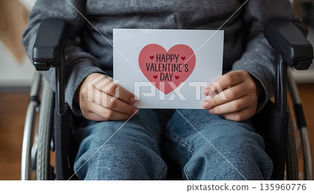 A disabled boy sits in a wheelchair and holds a Valentine's Day card. The concept is about Valentine's Day, inclusivity, love for others, and caring. 135960776